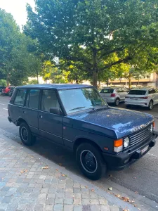 A somewhat old and ridiculously large black Range Rover parked in a street with lots of trees.