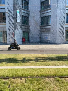 The top half of the picture is a grey building, in the center is the cobblestone pavement with a small asphalt road under it. On the left, we can see a slightly blurry person on a black scooter. The bottom half there is a patch of vivid green grass