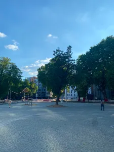 A cobble paved square shaded by trees with a couple people walking on it. The sky is blue, it's the evening.