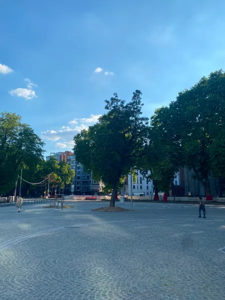 A cobble paved square shaded by trees with a couple people walking on it. The sky is blue, it's the evening.
