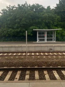 In the foreground you can see two sets of railtracks separated by a bit of rough gravel. In the background is an asphalt platform with a small shelter on the right and plenty of vegetation behind it.