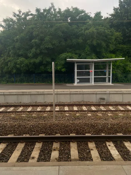 In the foreground you can see two sets of railtracks separated by a bit of rough gravel. In the background is an asphalt platform with a small shelter on the right and plenty of vegetation behind it.