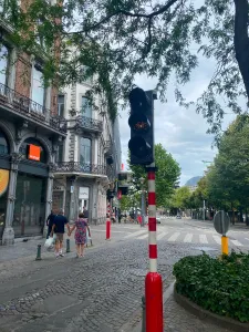 A picture of a slightly busy street. On the left you can see a couple of older people holding hands bringing back groceries and some more people further ahead. At the forefront in the center, you can see a tricolor bike lights post flashing orange. And on the right the street is lined with trees that block the view. The sky is cloudy and grayish and you can see a building with a dome far in the background.