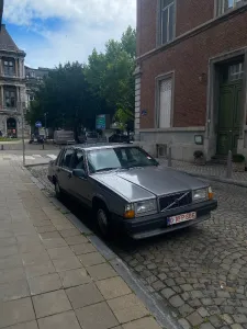 In the center of this image you can see and old boxy Volvo car parked on cobbled pavement. In the background there are trees and an old building with some signage.