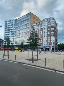 The subject of the picture is a set of three buildings separated by streets on each sides. The one on the left is white with loads of windows, the one in the middle is made out of concrete and has event more windows. They are both about 8 stories tall. The one on the right is in an older style, made out of red bricks and another white material. In the foreground, you can see a large pedestrian area with a couple people and a couple trees as well as a small one way road.