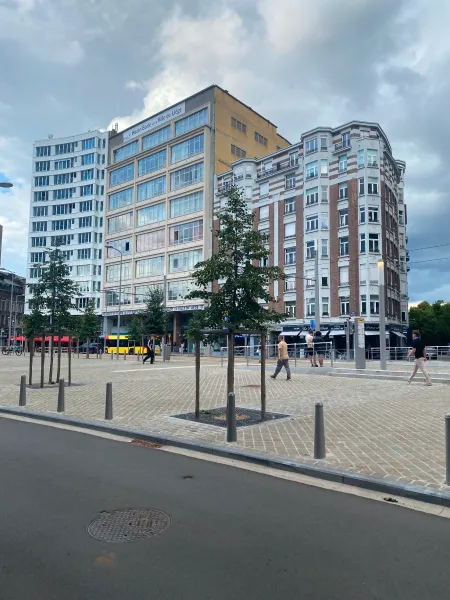 The subject of the picture is a set of three buildings separated by streets on each sides. The one on the left is white with loads of windows, the one in the middle is made out of concrete and has event more windows. They are both about 8 stories tall. The one on the right is in an older style, made out of red bricks and another white material. In the foreground, you can see a large pedestrian area with a couple people and a couple trees as well as a small one way road.