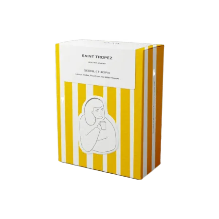 A yellow and white striped cardboard cofee box. On top is a white rectangular sticker that seals the box with Saint-Tropez written on it. On the front of the box is another sticker of a drawing of a person drinking a cup of coffee looking satisfied