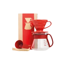 A red ceramic Hario V60 coffee dripper on top of it's glass carafe. There are also some filters laying down with a measuring spoon placed on top of them. In the back is the cardboard box in which it all comes.