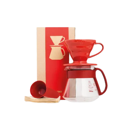 A red ceramic Hario V60 coffee dripper on top of it's glass carafe. There are also some filters laying down with a measuring spoon placed on top of them. In the back is the cardboard box in which it all comes.